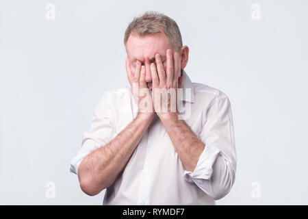 Senior man in white shirt covering his face with both hands Stock Photo