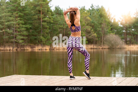 sporty woman standing on a jetty by a lake doing warm-up exercises. Back view. Stock Photo