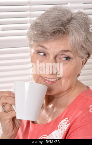 Happy elderly woman drinking tea on white window blinds background Stock Photo