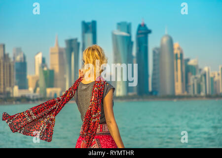 Back of caucasian woman looking at modern skyscrapers of Doha West Bay along Corniche promenade on Doha Bay in a sunny day. Female tourist visits Stock Photo