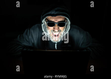 male hacker with beard and sunglasses working on his laptop, aggressive and potted expression, conceptual Stock Photo