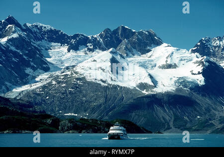 An excursion boat cruises past age-old snow-covered ice that slowly flows down rugged mountain valleys to Glacier Bay along the Inside Passage in southeast Alaska, USA. A ranger from Glacier Bay National Park and Preserve describes the awesome Alaskan terrain and wildlife seen on day-long tours that embarked from Bartlett Cove during the summer months. Besides icebergs and calving glaciers, passengers spot many seabirds and sea lions and may glimpse grizzly and black bears, moose, wolves and even mountain goats during the 130-mile (209-kilometer) water adventure in The Last Frontier. Stock Photo