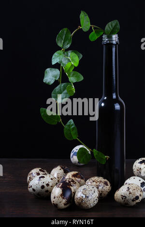 Quail eggs still life. Quail eggs lie on a dark wood table next to a green sprig in a black glass bottle on a black background Stock Photo