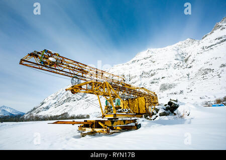Abandoned yellow metal construction or construction equipment on the background of snowy mountains and blue sky Stock Photo