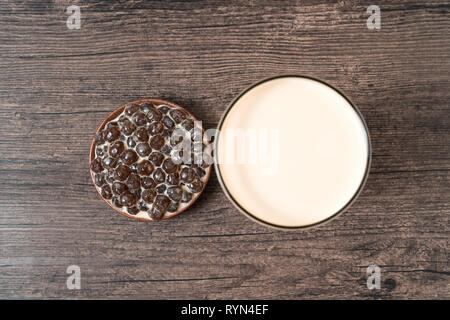 A glass cup of pearl milk tea (also called bubble tea) and a plate of tapioca ball on wooden background. Pearl milk tea is the most representative dri Stock Photo