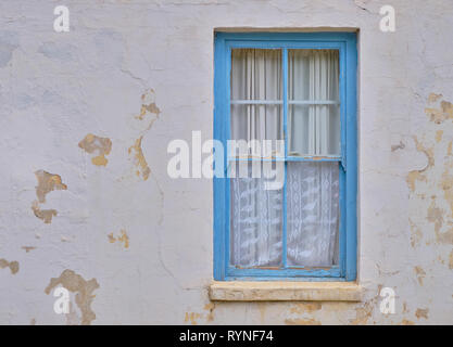 Minimalist capture of a single rural window on a white textured wall with peeling paint.  Simple window with blue frame, and white curtains Stock Photo