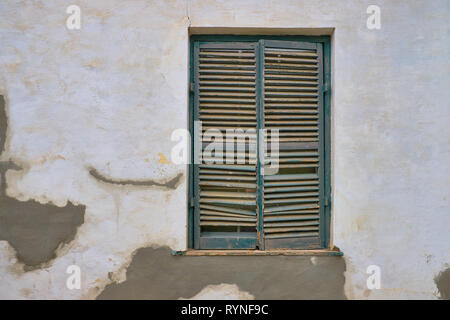 Minimalist capture of a single rural window on a white textured wall with plaster repairs.  Window has dark grey shutters in a state of needing repair Stock Photo