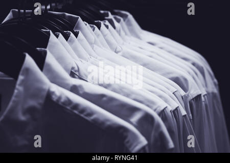 Row of white shirts hang on hangers in the darkness of a wardrobe, toned in blue. Stock Photo