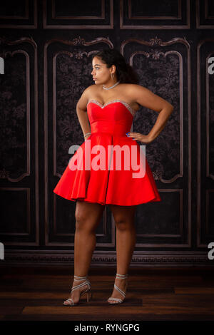 One beautiful,  biracial high school senior girl wearing red party dress waiting and looking away. Stock Photo