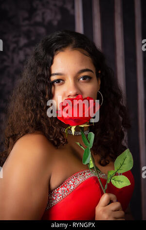 One beautiful,  biracial high school senior girl wearing red party dress holding one red rose Stock Photo