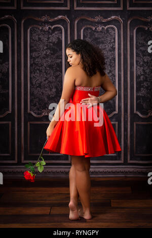One beautiful,  biracial high school senior girl wearing red party dress holding one red rose Stock Photo
