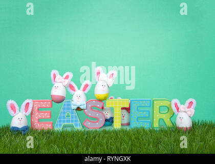 Colorful decorated Easter Eggs with bunny faces and ears sitting on and around decorative letter blocks spelling EASTER on grass with green background Stock Photo