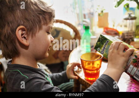 Little boy fills orange glass with cranberry juice from carton sitting at a table during healthy breakfast as parents teach him to be a help yourself. Stock Photo