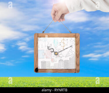 Hand holding whiteboard of dollar sign clock with business concept doodles on blue sky clouds green grass background. Time is money concept. Stock Photo