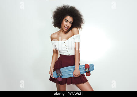 I love skating! Studio shot of beautiful young African woman in casual wear holding skateboard and smiling while standing against grey background. Active lifestyle. Sport concept Stock Photo