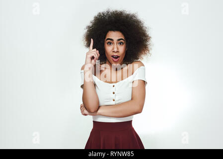 I have a great idea! Excited cute afro american woman is pointing her finger up and making a face with opened mouth while standing against grey background. Idea concept. Studio portrait Stock Photo
