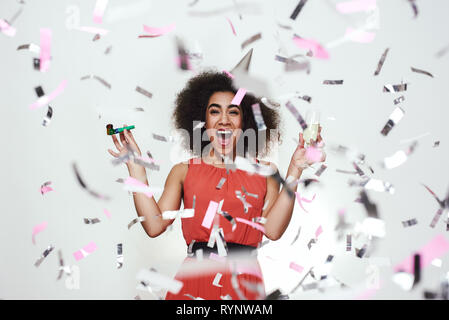 Portrait of a happy afro american woman in party hat holding glass of champagne and party whistle, while standing with opened mouth under confetti rain. Party concept. Happiness Stock Photo