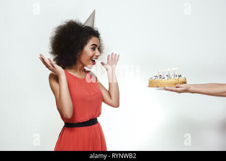 Surprise for you! Excited young afro american woman in party hat raising up her hands and looking at birthday cake with candles Stock Photo