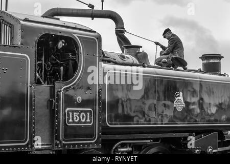 Black & white, close-up side view of vintage UK steam locomotive, stationary taking on water. Train driver, side view, on steam engine filling tank. Stock Photo