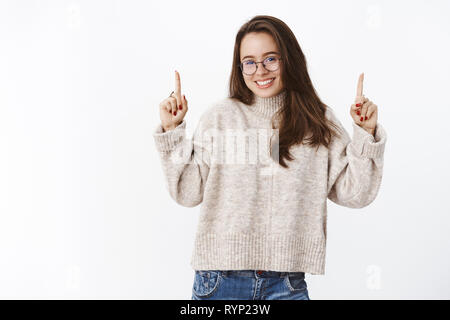 Waist-up shot of excited happy charming european girl in glasses and sweater raising hands pointing up and smiling from delight and joy as showing Stock Photo