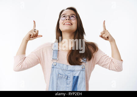 Waist-up shot of amused happy european female in glasses and overalls lifting head pointing and looking up mesmerized with fascinated and delighted Stock Photo
