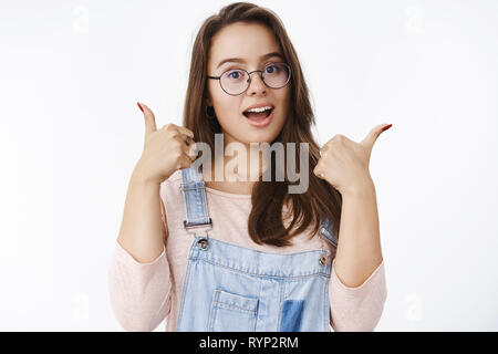 Waist-up shot of enthusiastic charming european female brunette in glasses and overalls showing thumbs up in like and approval, cheering for good Stock Photo
