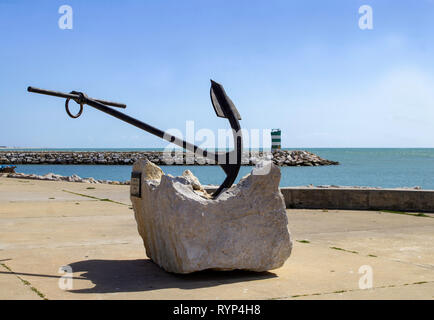Quarteira, Portugal. An old anchor mounted on a concrete base with a lighthouse in the background. Stock Photo