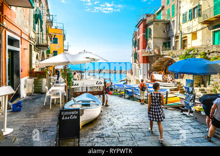 Tourists enjoy a sunny day on the Ligurian coast with the sea, cafe and boats in Riomaggiore Italy, on the Cinque Terre of the Italian Riviera Stock Photo