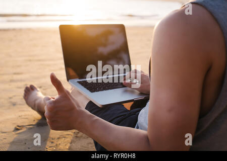 Close-up of young adult male traveler giving thumbs up and using computer laptop in casual summer clothes on tropical beach during sunset Stock Photo