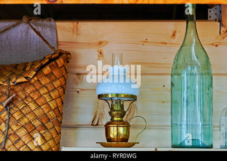 Still-life with an elegant vintage bronze oil lamp with white milk glass shade, wicker basket, and a glass bottle. Wooden wall in the background Stock Photo