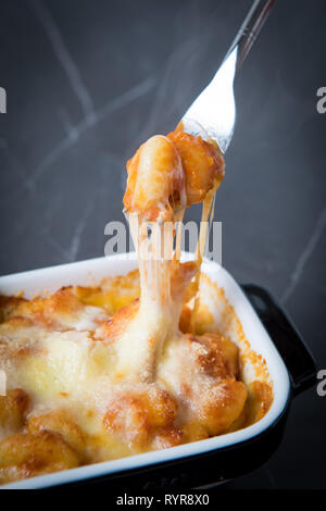 Oozing stringy cheese baked gnocchi pasta bake in a large baking tray. Fork pulling cheese in foreground over grey background. Gnocchi close up. Stock Photo