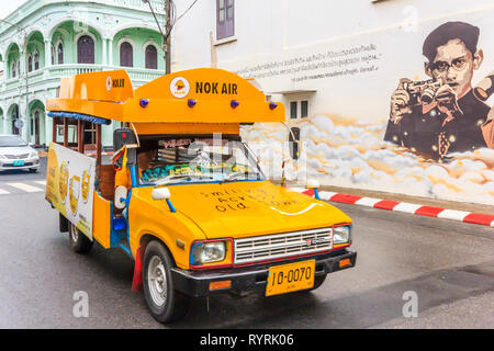 Phuket, Thailand - 11th April 2017: Yellow songthaew or taxi driving past mural of the late King. King Bhumibol was much revered. Stock Photo