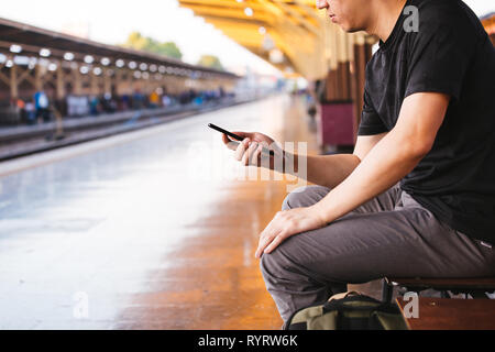Young male tourist and backpacker using a phone while waiting for a public transportation inside train platform Stock Photo