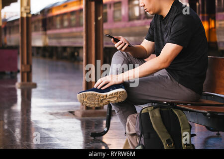 Young male tourist and backpacker using a phone while waiting for a public transportation inside train platform Stock Photo