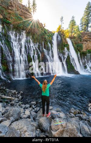 Young woman standing at a waterfall, stretching arms into the air, McArthur-Burney Falls Memorial State Park, California, USA Stock Photo