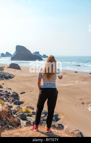Young woman looking over sandy beach to coast with rugged rocks, Myers Creek Beach Viewpoint, Oregon USA Stock Photo