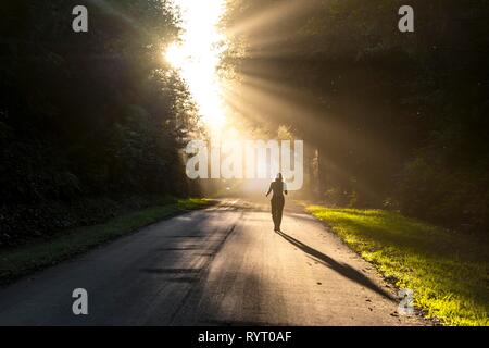 Young woman walking on a street, sunlight shining through trees, Oregon Coast Highway, Oregon, USA Stock Photo