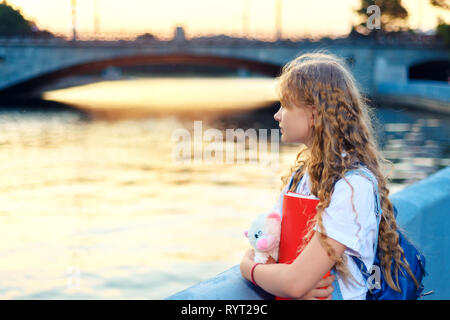 girl is standing near the river in the city at sunset. A teenager with a backpack is holding a bear toy and a red folder Stock Photo
