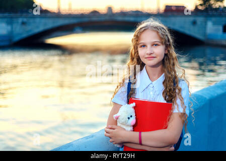 schoolgirl blonde with blue eyes is standing near the river in the city at sunset. teenager with holding a bear toy and a red folder Stock Photo