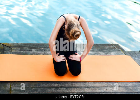 little girl doing yoga on the river bank. the child is engaged in pilates on an orange mat near the lake. healthy lifestyle Stock Photo