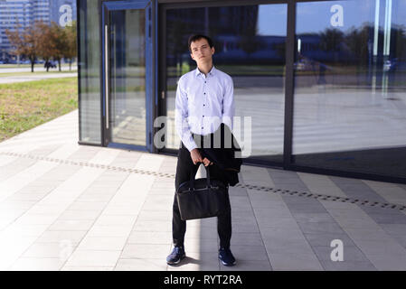 young businessman guy stands near building with large windows. man in shirt holds a briefcase and jacket in his hands Stock Photo