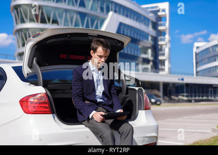 successful young businessman freelancer working outside in the city. man sit on car boot with tablet laptop Stock Photo
