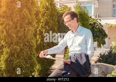young businessman in glasses reads documents and smiles in the street. the guy is holding jacket in his hands. toned Stock Photo