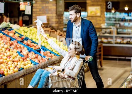 Man and woman having fun while riding in the cart with shopping list in the supermarket with fruits vegetables on the background Stock Photo