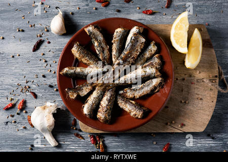 high angle view of some spanish grilled sardines in a brown earthenware plate, on a gray rustic wooden table sprinkled with some different spices, and Stock Photo