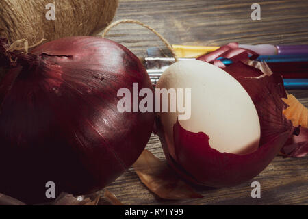 Fresh whole red onion, egg balancing in a dried onion skin and ball of brown twine on a wooden table. Making Easter eggs dyed with onion skins concept Stock Photo