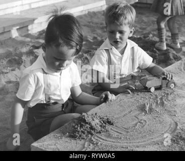 people, children, playing, two boys in the sandbox playing with sand and a toy truck, Germany, 1950s, children's playground, toy car, toy cars, car, cars, lorry, truck, toy, toys, game, fashion, clothes, male, manly, Germany, 20th century, children, child, kids, kid, boys, youngsters, sandbox, sandpit, sandboxes, sandpits, playing, play, historic, historical, half-length, half length, Additional-Rights-Clearance-Info-Not-Available Stock Photo