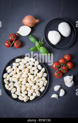 Gnocchi and mozarella on a black plate  and on a dark kitchen counter. Dark background. Ingredients for a baked gnocchi recipe. Tomato vines, garlic, Stock Photo