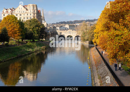 BATH, UK - OCTOBER 20, 2018: People walking by Pulteney Weir and Pulteney Bridge surrounded by vibrant autumnal trees on the River Avon in Bath, UK.