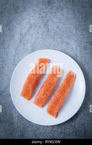 Three pieces of raw salmon with spices. Skin down pieces of fresh salmon on a white round plate and a grey background. Spiced healthy salmon filets un Stock Photo